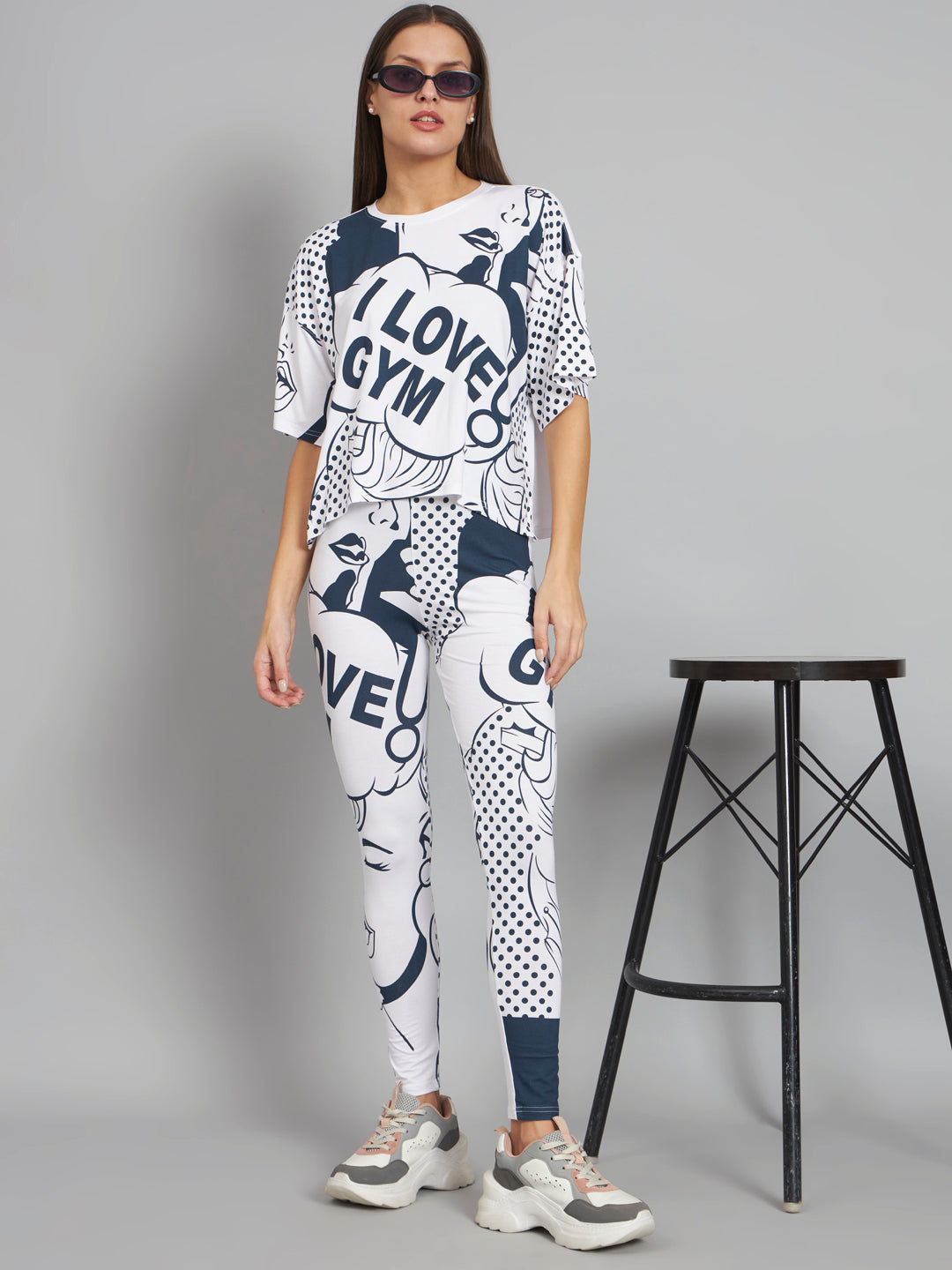 Woman wearing a white and blue patterned outfit with text, standing next to a black stool on a gray background.